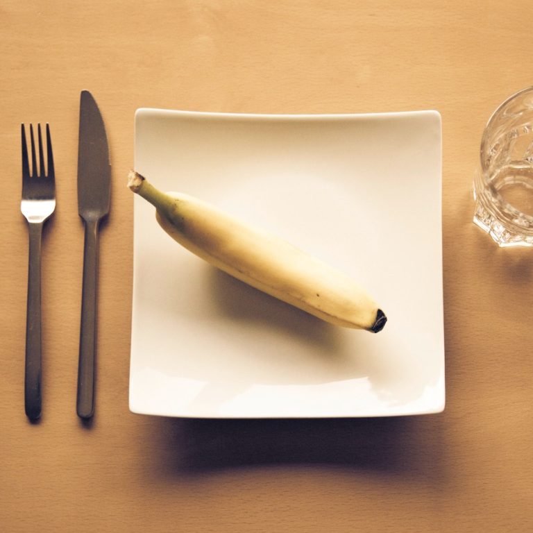 Eine Banane liegt auf einem eckigen, weißen Teller; links daneben liegt Besteck, rechts daneben steht ein Glas Wasser.